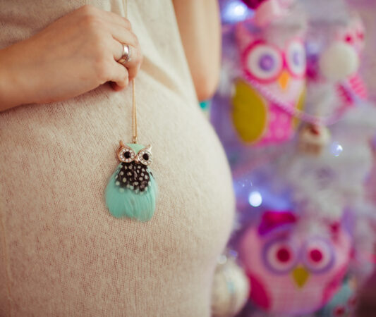 Femme enceinte portant un collier hibou turquoise de la collection baby dans un environnement coloré et joyeux