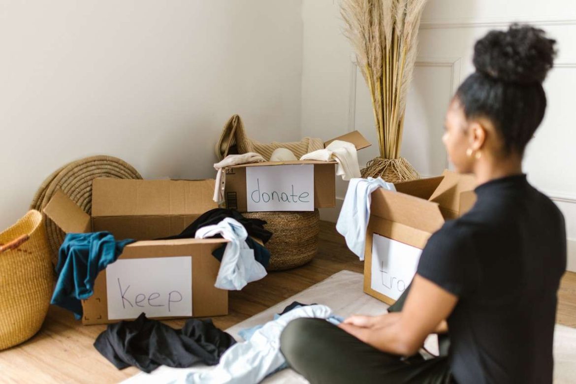 Une femme assise au sol applique la méthode pour faire le tri avant déménagement en répartissant ses vêtements dans des cartons étiquetés Garder, Donner et Jeter.